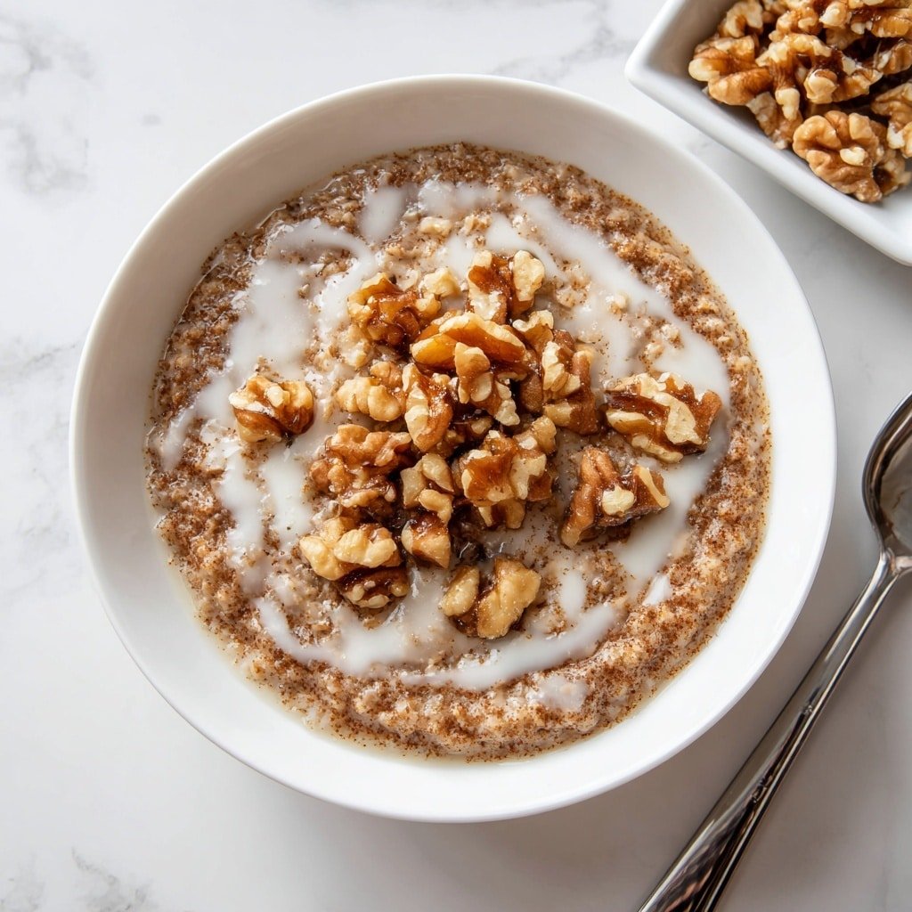 A round white bowl holds a thick, brown oatmeal base with a slightly rough texture. On top, there is a drizzle of white liquid in irregular patterns spread across the surface. In the center, a generous pile of chopped light brown walnuts covers part of the oatmeal, adding a chunky texture. The bowl sits on a white marbled surface, with a square white dish filled with more walnuts visible near the top right corner of the image. A silver spoon is partially shown on the right side. Photo taken with an iphone --ar 1:1 --v 7 — Creamy Spiced Red Split Lentil Porridge, nourishing breakfast recipes, comforting vegan dishes, coconut milk lentil porridge, hearty plant-based breakfast