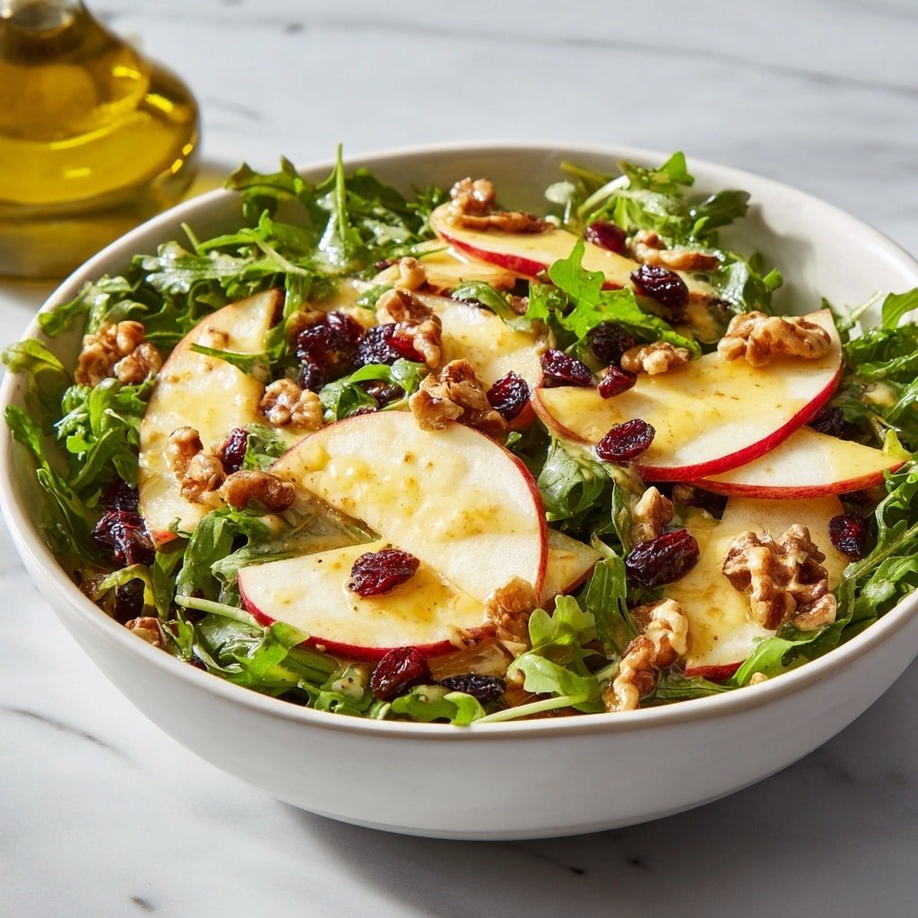 A white bowl filled with a fresh salad showing three main layers: the bottom layer is a bed of bright green leafy arugula with a slightly rough texture, the middle layer has thin, smooth, pale cream-colored apple slices with red edges, and the top layer is scattered with light brown walnut pieces and dark red dried cranberries. The salad is lightly drizzled with a golden dressing, creating a glossy look on the apple slices and greens. The bowl sits on a white marbled surface, and a blurred glass bottle of olive oil is visible in the background. Photo taken with an iphone --ar 1:1 --v 7 — Apple and Walnut Salad with Maple Dressing, apple walnut salad, maple dressing salad, fall fruit salad, healthy apple salad