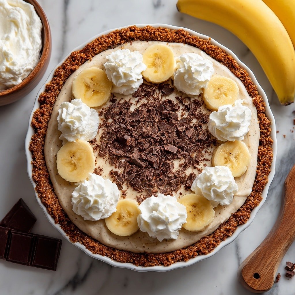 A round pie with a thick crumbly brown crust holds a creamy beige filling. On top, there are two visible layers of sliced bananas arranged in a ring near the outer edge. Between the banana slices and the center, dollops of white whipped cream are evenly placed. The center and the entire top are sprinkled with dark chocolate shavings, which add texture and color contrast. The pie sits on a white marbled surface, with pieces of dark chocolate and a yellow banana nearby. A wooden bowl with white cream is partially visible in the corner. photo taken with an iphone --ar 1:1 --v 7 — Black Bottom Banana Cream Pie, banana cream pie with chocolate layer, easy banana cream pie, decadent fruit and chocolate dessert, classic pie recipes